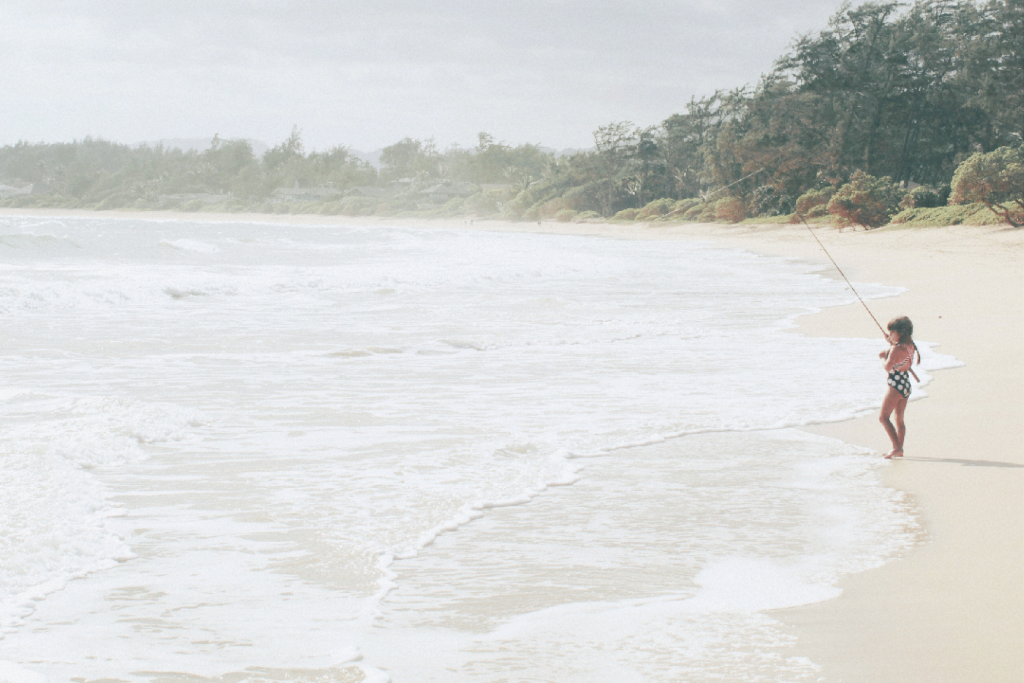 A long stretch of beach in Oahu with a nearby rainforest, where a small child is peacefully fishing—perfect for a relaxing Oahu itinerary
